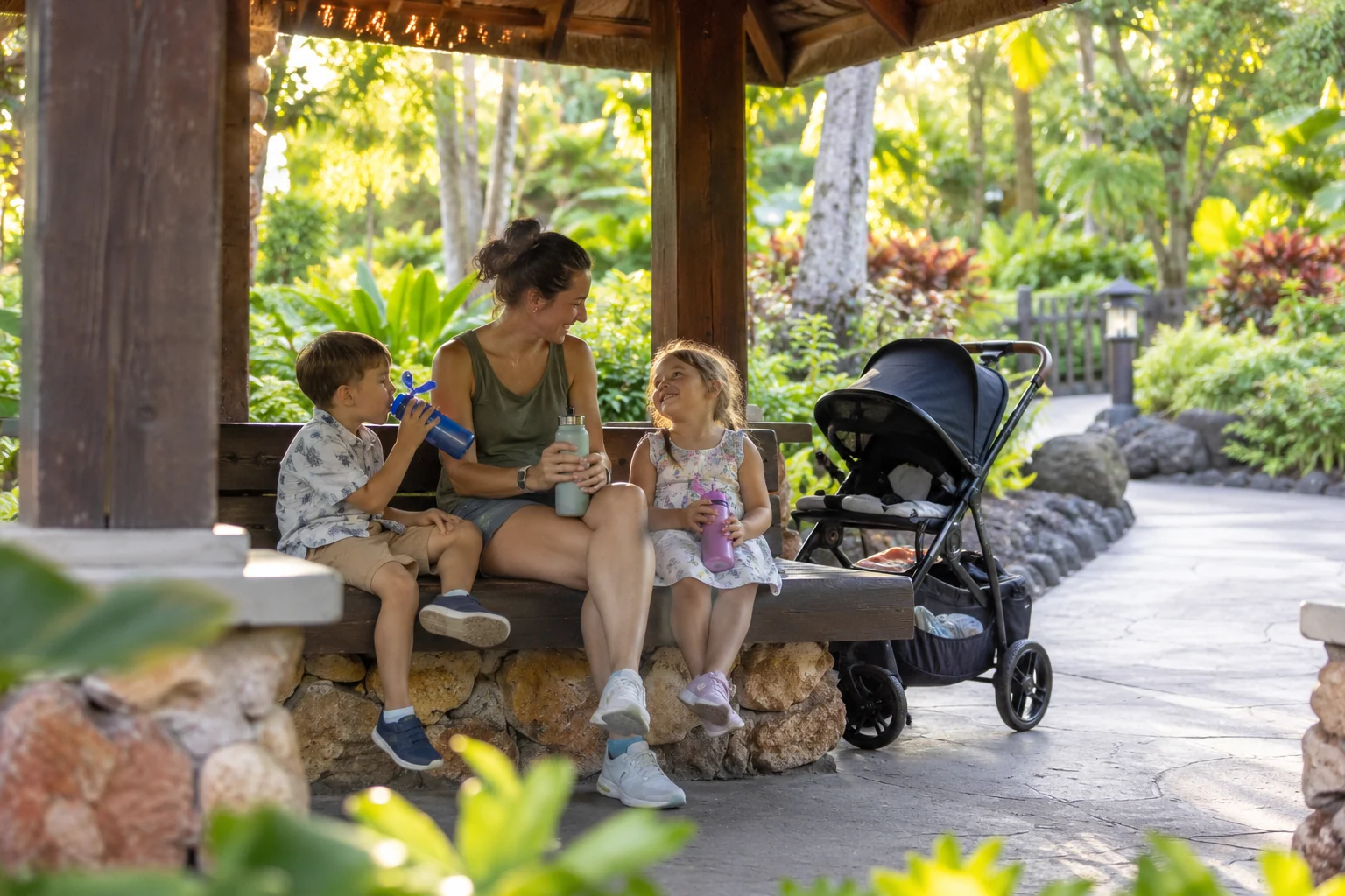 Parent and children taking a shaded rest and water break beside a tropical park path.
